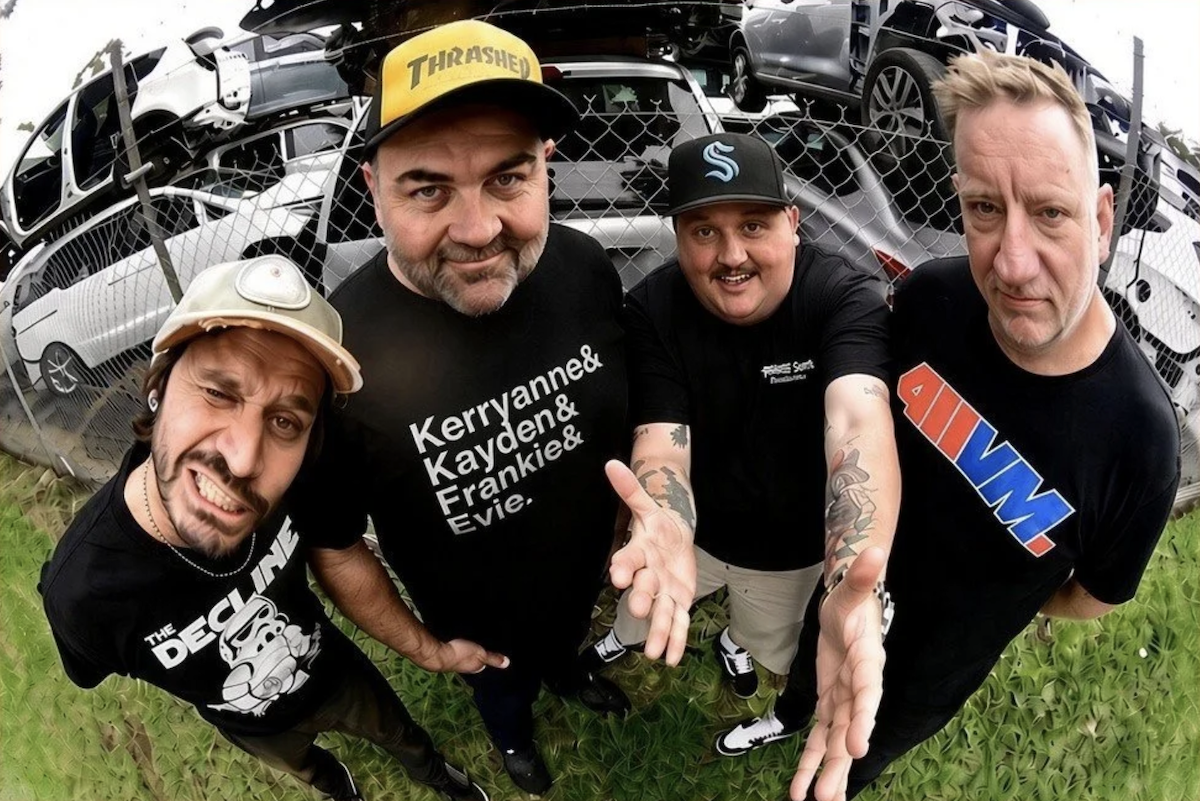 Punk rock band Not Dollar Short members posing outdoors in front of stacked cars at a scrapyard, captured with a fisheye wide-angle lens.